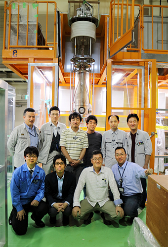 試験中の超伝導加速空洞（後ろ）と理化学研究所・高エネルギー加速器研究機構・三菱重工メカトロシステムズの研究開発グループの写真