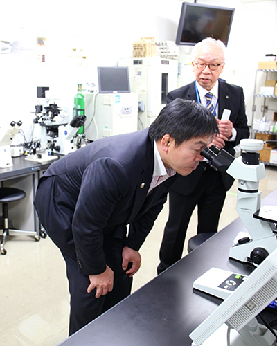 濱田博司センター長と顕微鏡を覗き込む馳浩（はせ ひろし）文部科学大臣