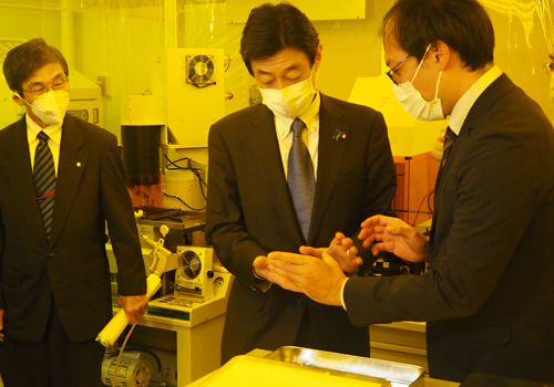 渡邉主任研究員（右）より説明を受けられる西村大臣（中央）の写真